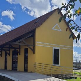 Imagem da fachada lateral do prédio histórico tombado pelo município de Itajaí conhecido como Estação Ferroviária Eng. Vereza que atualmente abriga as exposições do Museu Etno-Arqueológico de Itajaí. Sua arquitetura é composta por alvenaria e telhado em madeira, pelo sistema de mãos francesas. O prédio está printado na cor de amarelo ocre e sua fachada lateral possui detalhes arquitetônicos conhecidos como frisos e um elemento de triângulo. Também na imagem há uma parte da rampa que dá acesso as exposições.