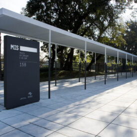 Entrada do Museu da Imagem e do Som (MIS), com uma ampla área pavimentada em primeiro plano. À esquerda, há um totem de cor preta com o nome do museu e o número 158. Ao lado, uma cobertura longa e branca, sustentada por pilares finos, forma um corredor que conduz até o edifício principal à direita. Ao fundo, vê-se uma área verde com árvores e grades, em um ambiente iluminado pela luz do dia.