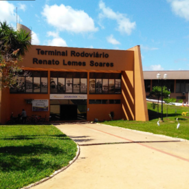 Fachada do Terminal Rodoviário Renato Lemes Soares em Dourados, onde está localizado o Museu Historico de Dourados com letreiro escuro no topo da entrada principal de cor terracota, cercado por gramado verde e palmeiras sob céu ensolarado.