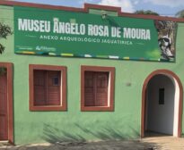 Fachada de prédio histórico pintado de verde, com duas janelas de madeira e uma porta em arco à direita. Acima da fachada há um grande banner verde com o nome “Museu Ângelo Rosa de Moura – Anexo Arqueológico Jaguatirica” e a imagem de uma jaguatirica.