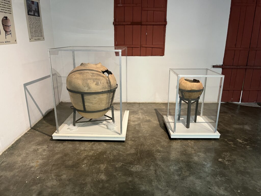 Duas urnas funerárias indígenas em cerâmica expostas em vitrines de vidro, apoiadas em suportes metálicos, em sala de exposição com piso de concreto e janelas de madeira ao fundo.