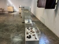 Sala de exposição com várias vitrines alinhadas contendo fragmentos cerâmicos e artefatos em pedra, em ambiente amplo com piso de concreto e janelas de madeira.