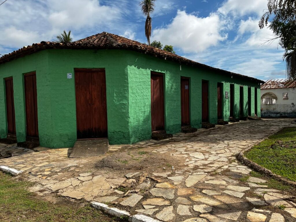 Prédio histórico verde com várias portas de madeira alinhadas, telhado de telhas cerâmicas e caminho de pedra ao longo da fachada, em área externa arborizada.