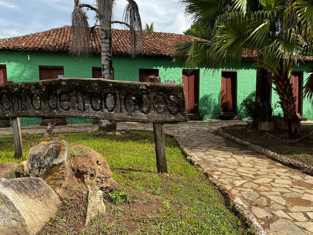 Placa de madeira com a inscrição “Centro de Tradições” em primeiro plano, com caminho de pedra e prédio histórico verde ao fundo, com portas de madeira e telhado colonial.