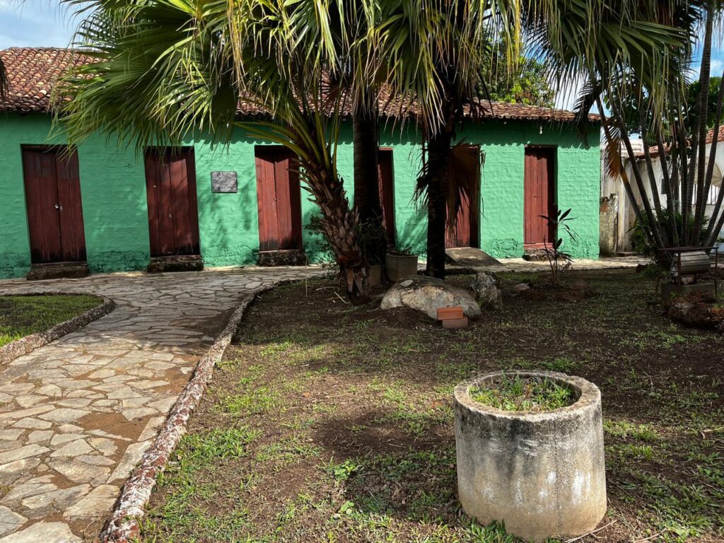 Jardim com palmeiras, caminho de pedra e prédio verde com várias portas de madeira, em área externa do Casarão histórico.