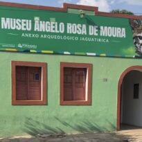 Fachada de prédio histórico pintado de verde, com duas janelas de madeira e uma porta em arco à direita. Acima da fachada há um grande banner verde com o nome “Museu Ângelo Rosa de Moura – Anexo Arqueológico Jaguatirica” e a imagem de uma jaguatirica.