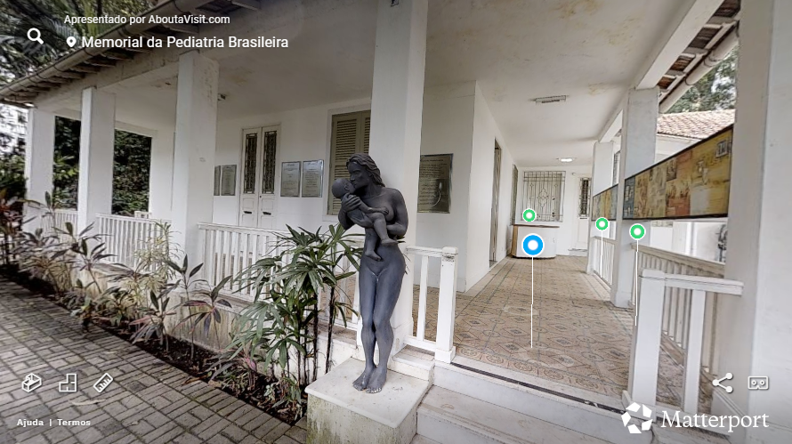 Imagem de um tour virtual no Memorial da Pediatria Brasileira. Em primeiro plano, há uma estátua em tom escuro representando uma mulher em pé segurando um bebê nos braços. A estátua está posicionada ao lado de uma pequena escada que leva a uma varanda coberta. A varanda possui piso decorado, colunas brancas e portas e janelas também brancas. Placas comemorativas estão fixadas na parede ao fundo. À direita, um corredor aberto segue ao longo da fachada, onde se vêem painéis expositivos afixados na parede. Plantas ornamentais ocupam a área ao lado da escada. O ambiente transmite a aparência de um espaço histórico e institucional.
