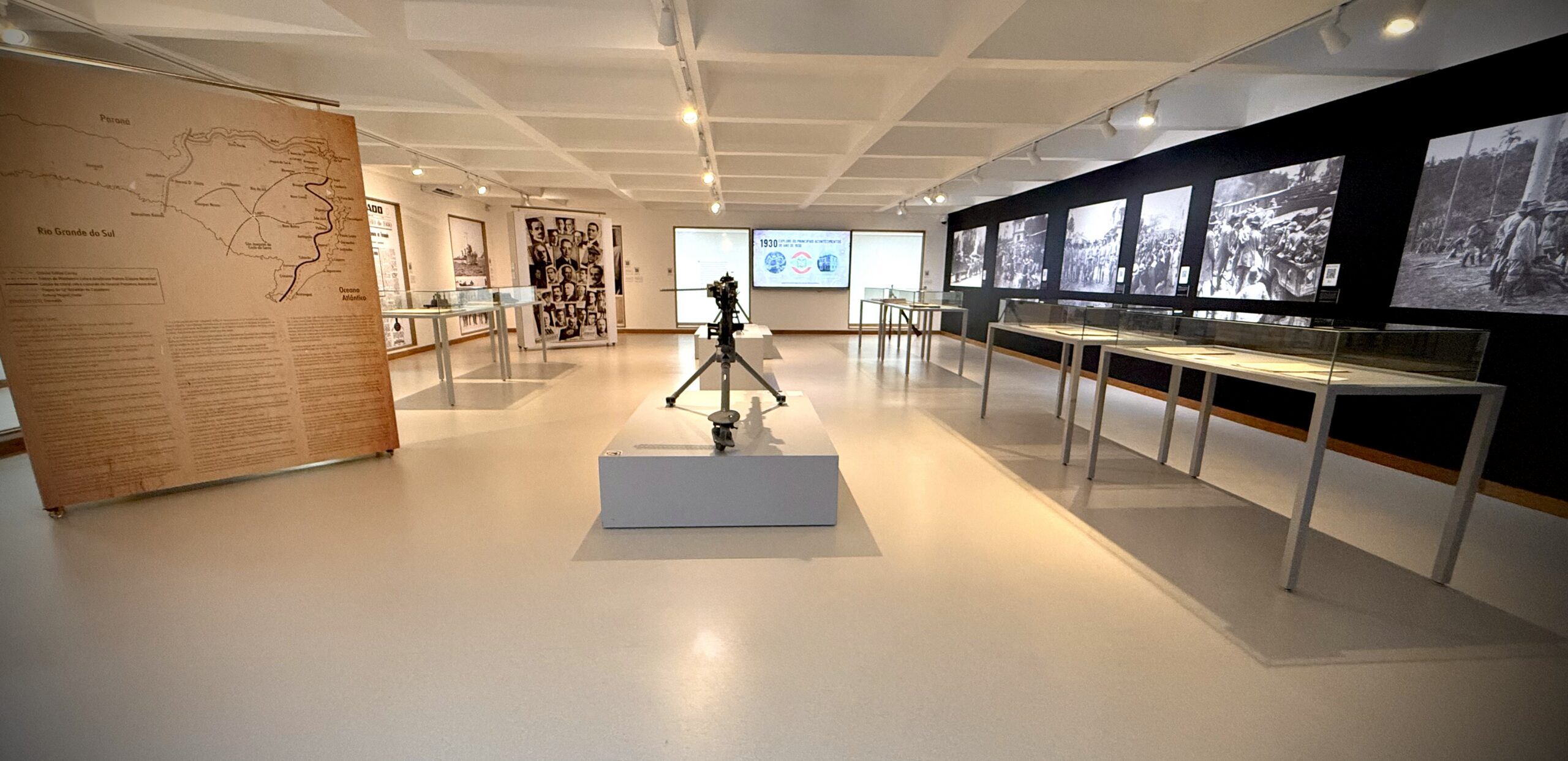 Vista ampla da sala de exposição. No centro, um objeto bélico antigo sobre pedestal. Ao redor, vitrines com documentos, painéis informativos e grandes fotografias históricas nas paredes laterais, sob iluminação uniforme e piso claro.