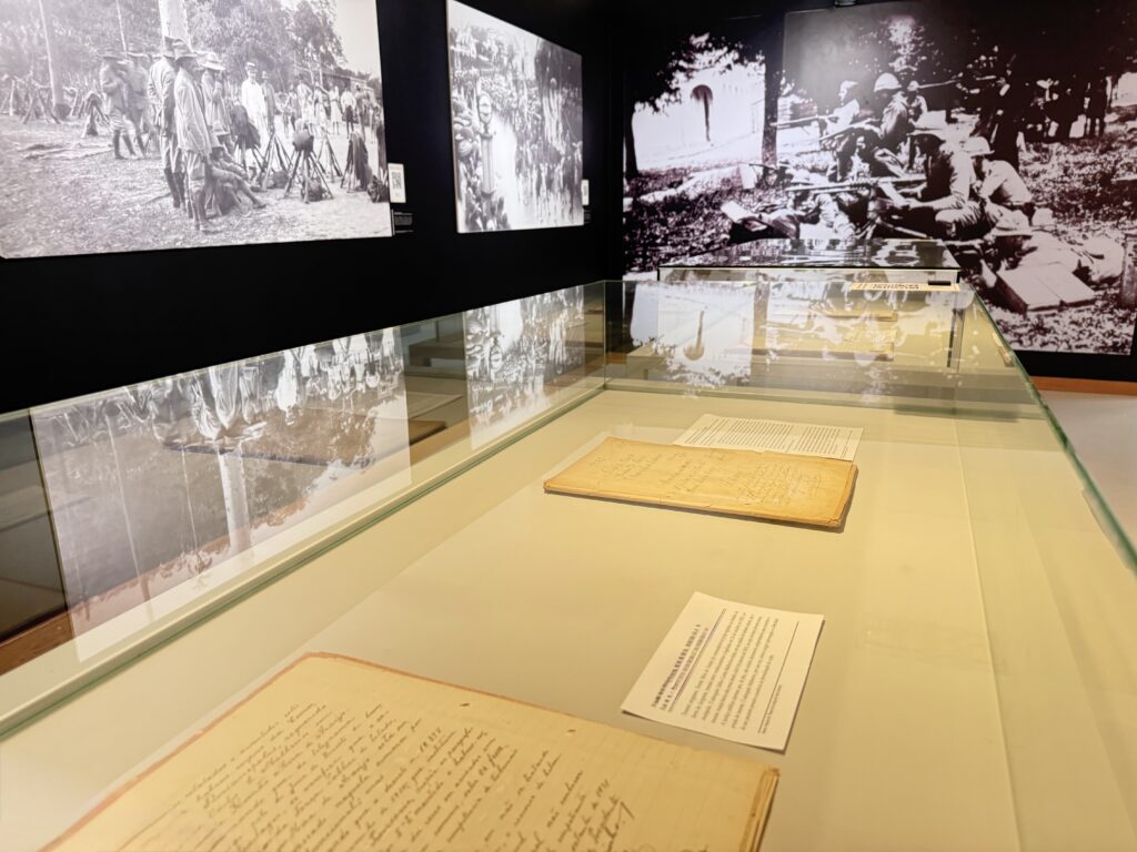 Vitrine horizontal contendo documentos manuscritos antigos. Ao fundo, grandes fotografias em preto e branco retratam cenas militares históricas, com soldados posicionados em campo aberto.