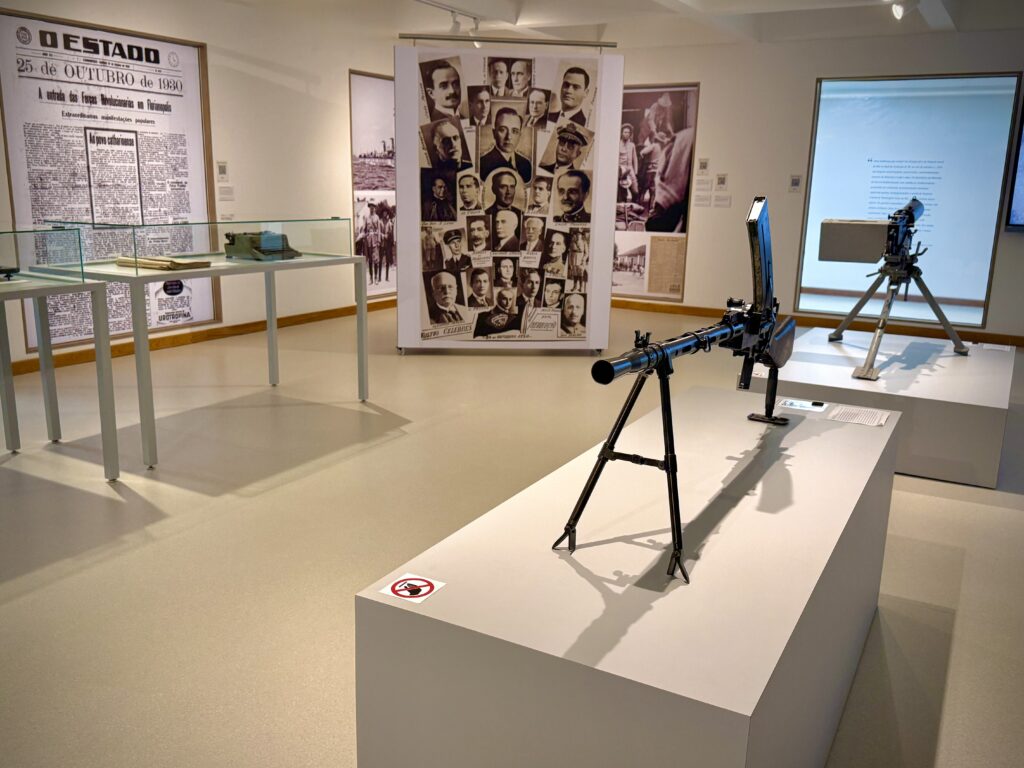 Sala expositiva com objetos históricos. Em primeiro plano, um equipamento bélico antigo montado sobre tripé, exposto em um pedestal branco. Ao fundo, vitrines com documentos, painéis fotográficos e reproduções de jornais históricos nas paredes.