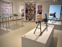 Sala expositiva com objetos históricos. Em primeiro plano, um equipamento bélico antigo montado sobre tripé, exposto em um pedestal branco. Ao fundo, vitrines com documentos, painéis fotográficos e reproduções de jornais históricos nas paredes.