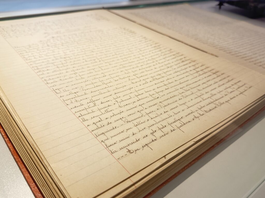 Livro manuscrito antigo aberto, exibido em vitrine horizontal de vidro. As páginas amareladas apresentam texto escrito à mão em tinta escura, com linhas regulares e margens visíveis.