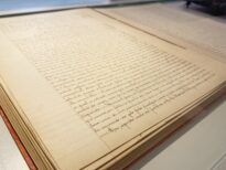 Livro manuscrito antigo aberto, exibido em vitrine horizontal de vidro. As páginas amareladas apresentam texto escrito à mão em tinta escura, com linhas regulares e margens visíveis.