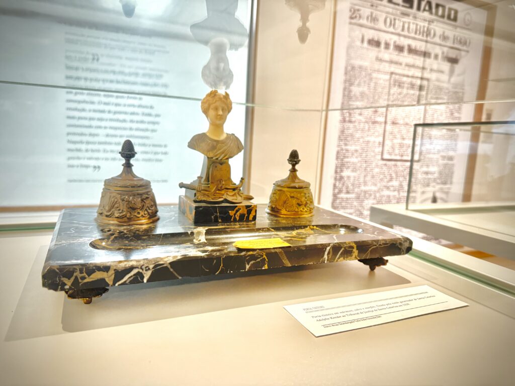 Na imagem aparece uma peça do acervo. Um objeto histórico exposto em vitrine de vidro: um tinteiro de mesa antigo em metal dourado, com três recipientes decorados e uma pequena escultura feminina ao centro, apoiados sobre uma base de mármore escuro com veios claros. Ao fundo, painéis com textos e reproduções de jornais históricos.