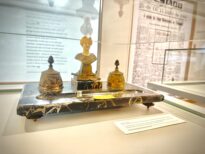 Na imagem aparece uma peça do acervo. Um objeto histórico exposto em vitrine de vidro: um tinteiro de mesa antigo em metal dourado, com três recipientes decorados e uma pequena escultura feminina ao centro, apoiados sobre uma base de mármore escuro com veios claros. Ao fundo, painéis com textos e reproduções de jornais históricos.