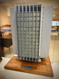 Maquete arquitetônica em tons de cinza representando um edifício moderno, com fachada marcada por brises verticais. A maquete está sobre uma base de madeira com placa identificando o Palácio da Justiça Ministro Luiz Gallotti.