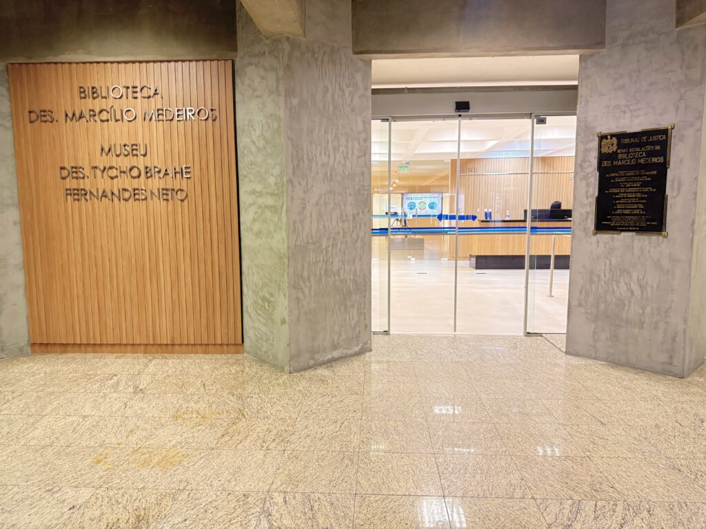 Entrada do Museu Tycho Brahe Fernandes Neto e da Biblioteca. Na imagem estão paredes de concreto aparente e piso de granito claro. À esquerda, um painel de madeira com a inscrição “Biblioteca Des. Marcilio Medeiros” e “Museu Des. Tycho Brahe Fernandes Neto”. Ao centro, portas de vidro dão acesso ao interior do museu. À direita, uma placa institucional em metal fixada na parede.