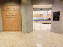 Entrada do Museu Tycho Brahe Fernandes Neto e da Biblioteca. Na imagem estão paredes de concreto aparente e piso de granito claro. À esquerda, um painel de madeira com a inscrição “Biblioteca Des. Marcilio Medeiros” e “Museu Des. Tycho Brahe Fernandes Neto”. Ao centro, portas de vidro dão acesso ao interior do museu. À direita, uma placa institucional em metal fixada na parede.