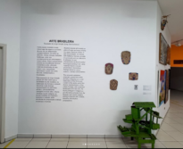 Parede branca da sala de exposição permanente do MAPA, onde há um breve texto sobre arte brasileira, além de obras em argila fixadas na parece e uma obra em madeira e lata na cor verde mais próxima ao chão