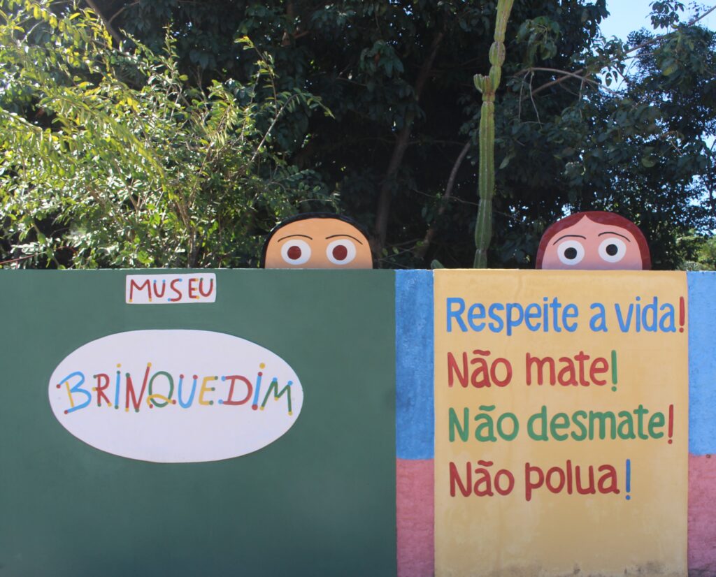 o Descrição Geral: A imagem mostra um muro colorido, cercado por uma vegetação densa de árvores verdes e um cacto alto ao fundo, sob a luz do sol. O muro é dividido em seções de cores vibrantes e por cima se vê cabeças de bonecos que olham para o lado de fora. O da esquerda tem cabelos pretos e o da direita cabelos avermelhados. Ambos possuem olhos brancos muito grandes e redondos, com pupilas pretas, criando uma expressão de curiosidade e surpresa. Detalhes da Composição: Do lado Esquerdo a parede é pintada de verde escuro. No topo, há uma pequena placa retangular branca com a palavra "MUSEU" em letras vermelhas. Logo abaixo, há um grande oval branco que contém o nome "BRINQUEDIM". As letras são estilizadas, coloridas (azul, amarelo, vermelho e verde) e possuem pequenos pontos nas extremidades. Do lado Direito tem um Mural de Mensagens o Esta seção tem um fundo amarelo claro, com bordas laterais em azul e uma base rosa. o Nela, está escrita a seguinte mensagem em letras grandes, cada linha em uma cor diferente:  Respeite a vida! (em azul)  Não mate! (em vermelho)  Não desmate! (em verde)  Não polua! (em marrom/bordô)