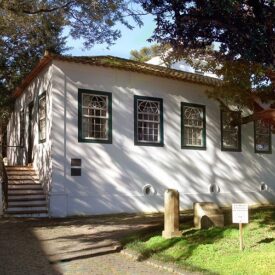 Em um dia ensolarado, a fachada do Museu. O casarão tem telhas de barro, paredes brancas e as molduras das janelas são verdes. Na lateral, uma escadaria dá acesso à porta principal. entorno, muitas árvores.