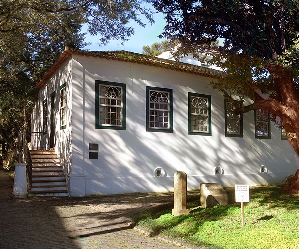 Em um dia ensolarado, a fachada do Museu. O casarão tem telhas de barro, paredes brancas e as molduras das janelas são verdes. Na lateral, uma escadaria dá acesso à porta principal. entorno, muitas árvores.