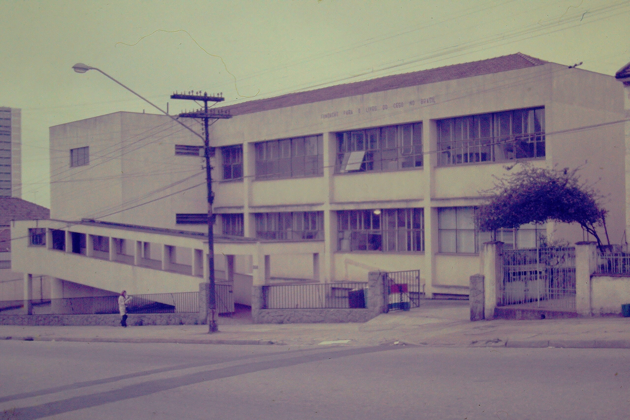 Fotografia da fachada da Fundação Dorina Nowill para Cegos. O edifício é de dois pavimentos, com arquitetura simples e linhas retas, pintura clara e grandes janelas. Na parte superior da fachada, há a inscrição com o nome da instituição "Fundação para o Livro do Cego no Brasil/'. À frente do prédio, vê-se uma rampa de acesso, um portão e a calçada junto à rua, com poste de iluminação e fiação aérea, indicando o contexto urbano da época.