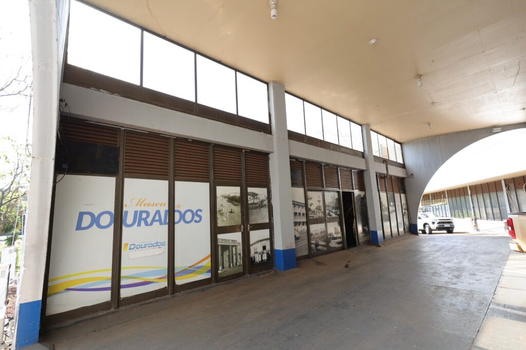 Fotografia em ângulo diagonal da fachada de entrada do Museu Histórico e Cultural de Dourados. A entrada fica sob uma grande cobertura de concreto e é composta por várias portas de vidro sequenciais com esquadrias escuras. Os vidros estão envelopados com adesivos: à esquerda, lê-se "Museu de Dourados" em letras azuis e amarelas; à direita, há reproduções de fotografias antigas em preto e branco que mostram a história do município.
