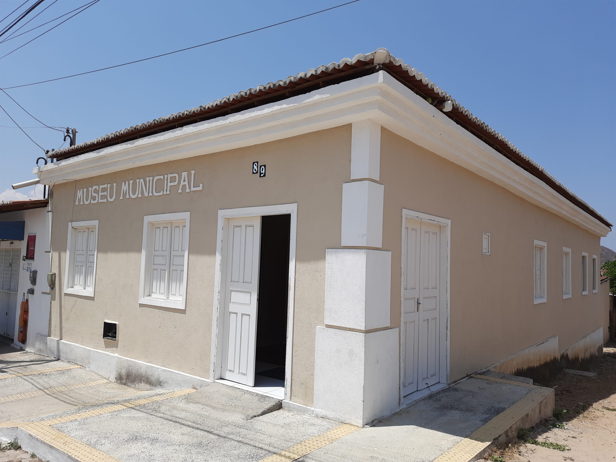 Fachada do Museu Municipal em um prédio térreo de cor bege clara, com detalhes brancos nas portas, janelas e colunas. A entrada principal está aberta, com uma porta dupla branca, acima da qual se lê "MUSEU MUNICIPAL" em letras grandes. O telhado de cerâmica vermelha contrasta com o céu azul claro e sem nuvens. A calçada em frente possui piso tátil para acessibilidade.