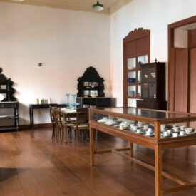 Fotografia de uma sala de exposição do museu. O espaço possui piso de madeira e está mobiliado com vitrines e móveis antigos em madeira escura, incluindo cristaleiras, armários e uma mesa com cadeiras. Objetos como louças, copos e peças decorativas estão dispostos nas vitrines e prateleiras. A parede branca contrasta com as portas e janelas em tom avermelhado. Um extintor de incêndio está fixado ao lado da porta à direita.
