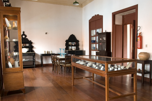 Fotografia de uma sala de exposição do museu. O espaço possui piso de madeira e está mobiliado com vitrines e móveis antigos em madeira escura, incluindo cristaleiras, armários e uma mesa com cadeiras. Objetos como louças, copos e peças decorativas estão dispostos nas vitrines e prateleiras. A parede branca contrasta com as portas e janelas em tom avermelhado. Um extintor de incêndio está fixado ao lado da porta à direita.
