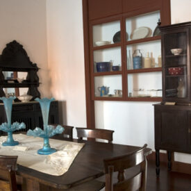 Fotografia da sala expositiva do museu. O ambiente apresenta uma mesa retangular de madeira com cadeiras ao redor e candelabros de vidro azul sobre uma toalha clara. Ao fundo, há móveis antigos, como cristaleiras e aparadores, que expõem louças e utensílios. Na parede, uma vitrine embutida exibe diversas peças em cerâmica e vidro. O piso é de madeira e as paredes brancas contrastam com os elementos em madeira escura.