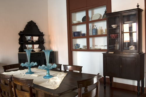 Fotografia da sala expositiva do museu. O ambiente apresenta uma mesa retangular de madeira com cadeiras ao redor e candelabros de vidro azul sobre uma toalha clara. Ao fundo, há móveis antigos, como cristaleiras e aparadores, que expõem louças e utensílios. Na parede, uma vitrine embutida exibe diversas peças em cerâmica e vidro. O piso é de madeira e as paredes brancas contrastam com os elementos em madeira escura.