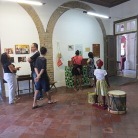 Fotografia de sala de exposição com piso de ladrilho cerâmico e paredes brancas, marcada por arcos de tijolos aparentes. Diversas pessoas observam painéis informativos, objetos pendurados e imagens expostas na parede. Em primeiro plano, uma criança vestida com roupa tradicional está próxima a dois tambores. A iluminação é feita por luminárias suspensas.