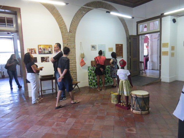 Fotografia de sala de exposição com piso de ladrilho cerâmico e paredes brancas, marcada por arcos de tijolos aparentes. Diversas pessoas observam painéis informativos, objetos pendurados e imagens expostas na parede. Em primeiro plano, uma criança vestida com roupa tradicional está próxima a dois tambores. A iluminação é feita por luminárias suspensas.