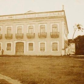 Imagem histórica em tons de sépia da fachada do Museu. O edifício possui dois andares, revestido na fachada com azulejos. A foto está recortada e no térreo é possível observar quatro janelas e uma porta principal mais ampla, dispostas de forma ritmada. No andar superior, nota-se cinco portas, todas com grades de ferro. Em frente ao edifício, existe um espaço verde.