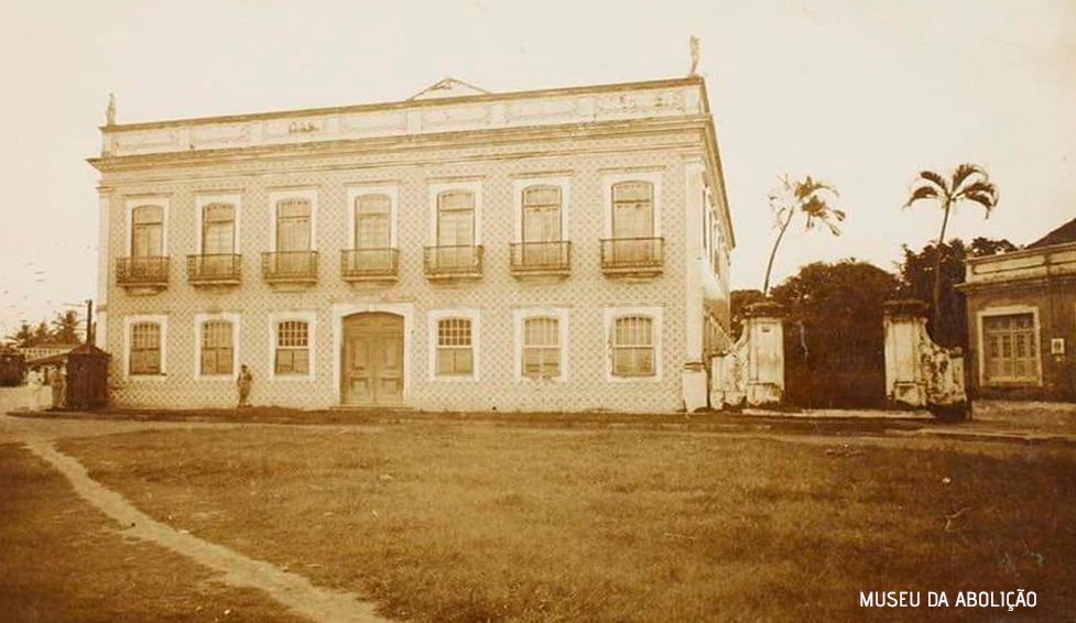 Imagem histórica em tons de sépia da fachada do Museu. O edifício possui dois andares, revestido na fachada com azulejos. A foto está recortada e no térreo é possível observar quatro janelas e uma porta principal mais ampla, dispostas de forma ritmada. No andar superior, nota-se cinco portas, todas com grades de ferro. Em frente ao edifício, existe um espaço verde.