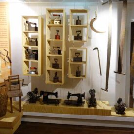Fotografia de uma das salas da exposição de longa duração do museu. No centro, encostada na parede, há três prateleiras de madeira com diversos ferros de passar roupa antigos. Na parte inferior do móvel, estão expostas várias máquinas de costura antigas. À direita, penduradas na parede, uma foice e duas serras grandes. À esquerda, há um cesto de vime e um banco antigo ao lado de um painel com texto explicativo. No canto inferior direito do piso da sala, há um extintor de incêndio vermelho.