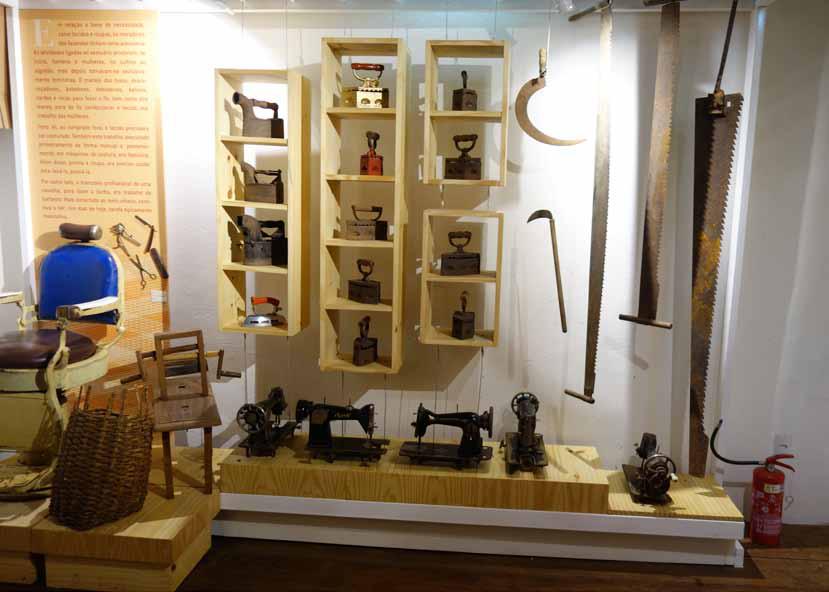 Fotografia de uma das salas da exposição de longa duração do museu. No centro, encostada na parede, há três prateleiras de madeira com diversos ferros de passar roupa antigos. Na parte inferior do móvel, estão expostas várias máquinas de costura antigas. À direita, penduradas na parede, uma foice e duas serras grandes. À esquerda, há um cesto de vime e um banco antigo ao lado de um painel com texto explicativo. No canto inferior direito do piso da sala, há um extintor de incêndio vermelho.