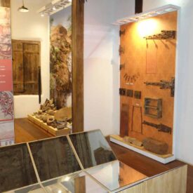 Fotografia de uma sala de exposição do museu. À esquerda, nota-se uma parede com painéis informativos e um portal de madeira que leva a outro ambiente. À direita, uma parede exibe ferramentas antigas e outros artefatos, com iluminação direcionada destacando esses itens. No ambiente ao fundo, há uma janela de madeira e na parede à direita estão expostos alguns utensílios. O piso da sala é de madeira e as paredes estão pintadas de branco.