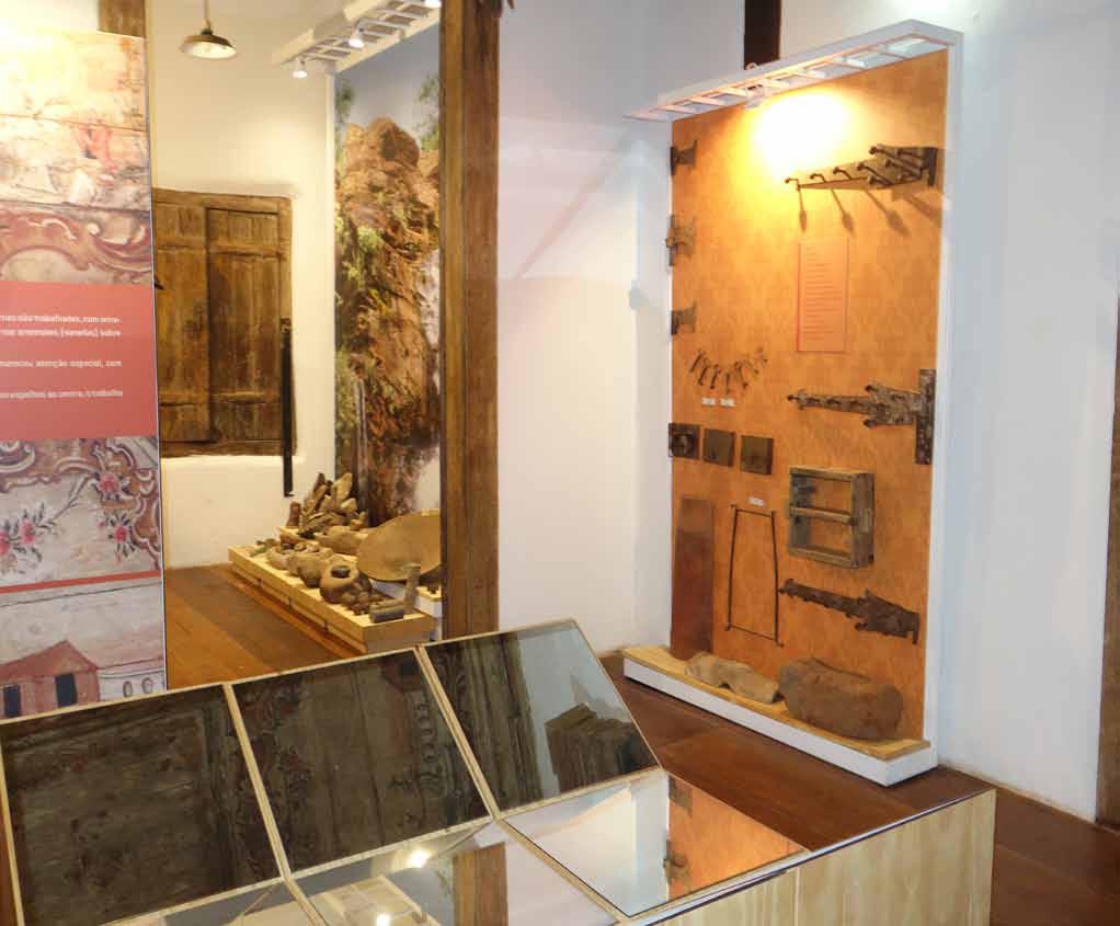 Fotografia de uma sala de exposição do museu. À esquerda, nota-se uma parede com painéis informativos e um portal de madeira que leva a outro ambiente. À direita, uma parede exibe ferramentas antigas e outros artefatos, com iluminação direcionada destacando esses itens. No ambiente ao fundo, há uma janela de madeira e na parede à direita estão expostos alguns utensílios. O piso da sala é de madeira e as paredes estão pintadas de branco.