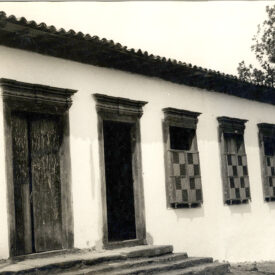 Fotografia histórica em preto e branco do museu. À esquerda, há uma porta de madeira; no centro, uma porta aberta; e à direita, duas janelas com treliças de madeira dispostas em padrão quadriculado. A cobertura é de telhas, com beiral estreito, e há degraus que permitem acessar o Museu.