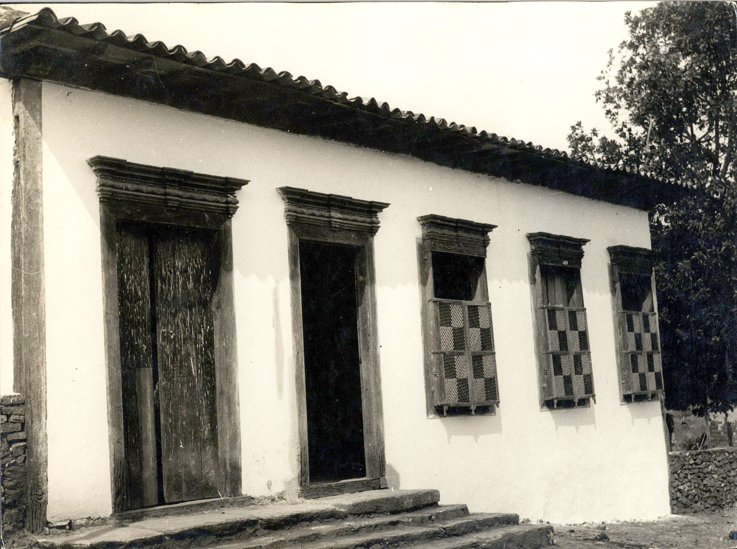 Fotografia histórica em preto e branco do museu. À esquerda, há uma porta de madeira; no centro, uma porta aberta; e à direita, duas janelas com treliças de madeira dispostas em padrão quadriculado. A cobertura é de telhas, com beiral estreito, e há degraus que permitem acessar o Museu.