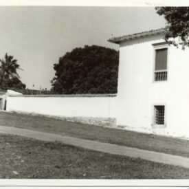 Fotografia histórica em preto e branco da fachada lateral do museu com parede externa branca. A imagem mostra a fachada da construção com duas janelas: uma no andar superior com grades e outra no térreo. Ao lado do edifício, há um muro alto que se estende ao fundo da imagem. Atrás do muro, é possível ver árvores. Em primeiro plano, há um caminho pavimentado que corre paralelo ao prédio e ao muro.