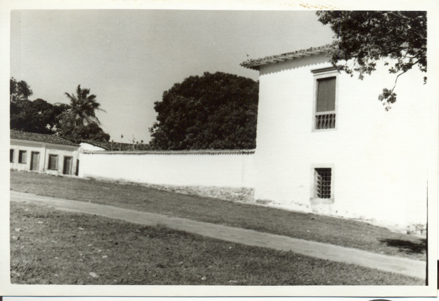 Fotografia histórica em preto e branco da fachada lateral do museu com parede externa branca. A imagem mostra a fachada da construção com duas janelas: uma no andar superior com grades e outra no térreo. Ao lado do edifício, há um muro alto que se estende ao fundo da imagem. Atrás do muro, é possível ver árvores. Em primeiro plano, há um caminho pavimentado que corre paralelo ao prédio e ao muro.