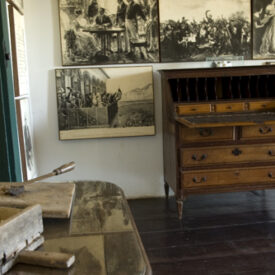 Imagem de sala expositiva com vários quadros na parede e uma escrivaninha antiga de madeira, com gavetas e uma tampa aberta. Na outra parede, há uma mesa com instrumentos que parecem ferramentas de trabalho. A sala tem paredes brancas e piso de madeira escura, com iluminação natural.