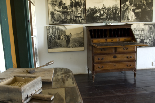 Imagem de sala expositiva com vários quadros na parede e uma escrivaninha antiga de madeira, com gavetas e uma tampa aberta. Na outra parede, há uma mesa com instrumentos que parecem ferramentas de trabalho. A sala tem paredes brancas e piso de madeira escura, com iluminação natural.