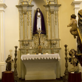 Fotografia de um altar, onde estátuas de figuras religiosas, como a Virgem Maria e o Menino Jesus, são o foco. Os detalhes de suas roupas e poses demonstram o trabalho artesanal e a busca pela representação realista das figuras.