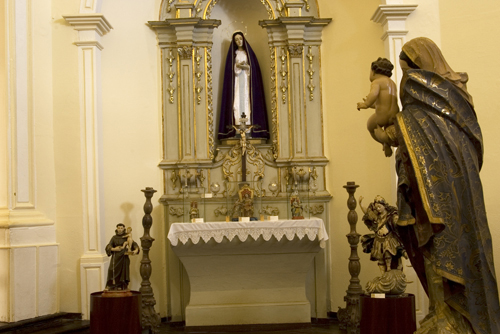 Fotografia de um altar, onde estátuas de figuras religiosas, como a Virgem Maria e o Menino Jesus, são o foco. Os detalhes de suas roupas e poses demonstram o trabalho artesanal e a busca pela representação realista das figuras.