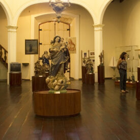 Fotografia da principal sala de exposição do Museu, com piso de madeira escura e colunas com arcos. Estão expostas esculturas sacras sobre bases de madeira, com destaque, ao centro, para uma grande imagem feminina com anjos aos seus pés. Há visitantes observando as obras e, no fundo da sala, uma escada de madeira à esquerda.